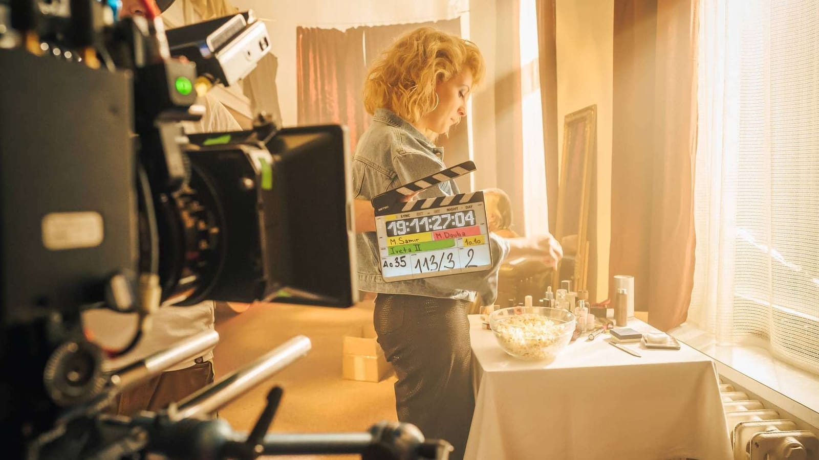 Woman holding a clapperboard on the set of the Voyo series Iveta, camera in the foreground and a sunlit window behind