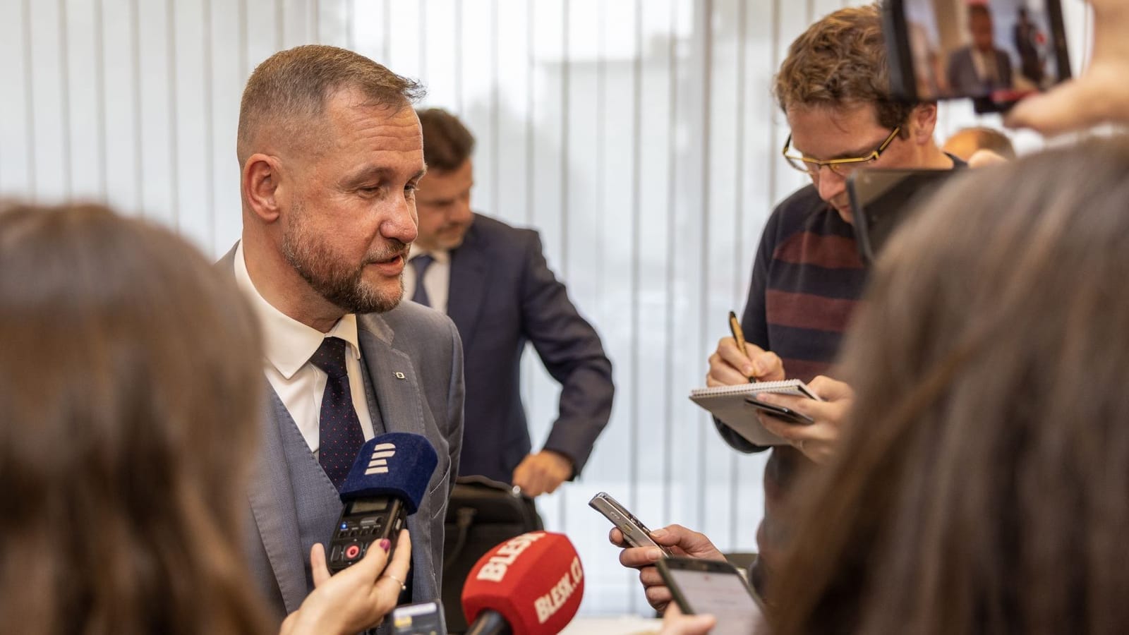 Jan Souček speaking to reporters holding microphones labelled ČT and Blesk.cz as a journalist takes notes nearby