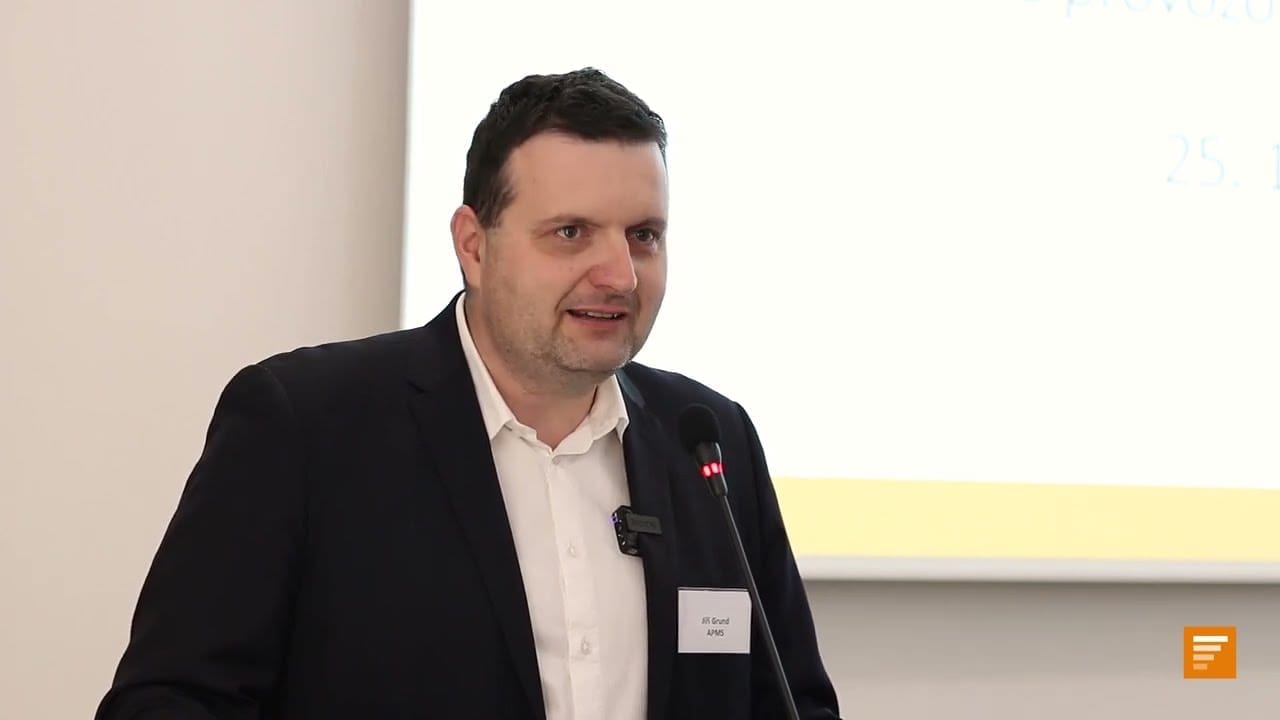 Jiří Grund in a dark suit with a name badge speaking at a microphone in front of a projection screen