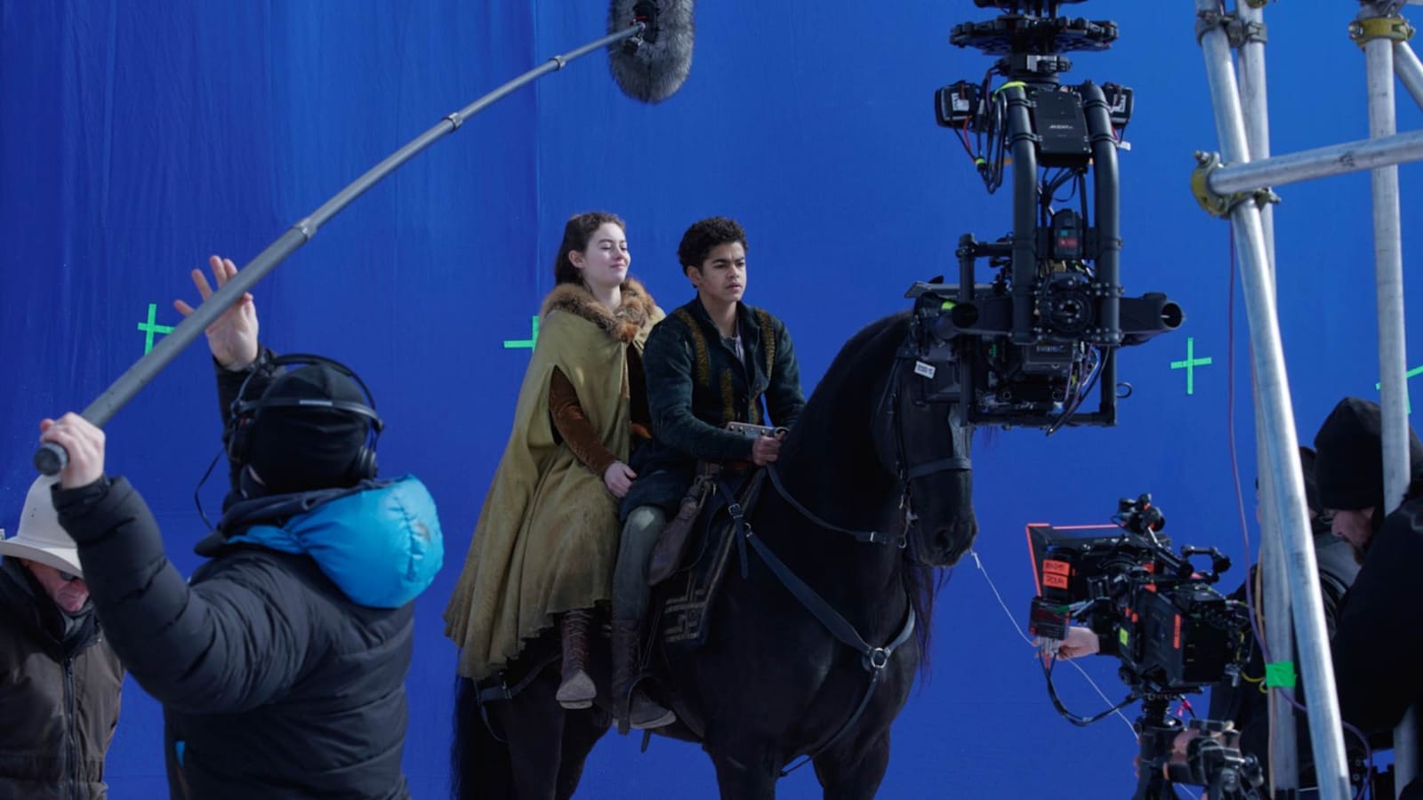 Two young actors in period costume ride a black horse on a film set in front of a blue screen, surrounded by a boom operator, camera crane and crew