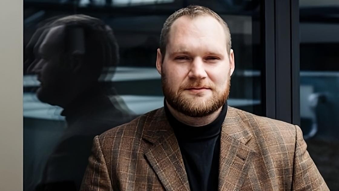 Tomáš Vojáček, a bearded man in a brown checked jacket and black turtleneck, standing by a glass wall