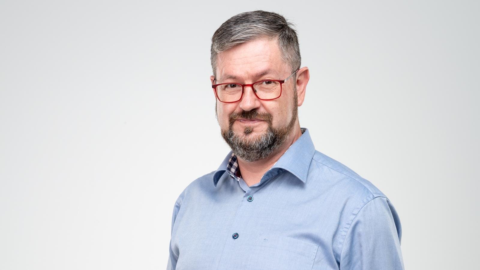 Pavel Novák, a bearded man with grey hair and red glasses in a light blue shirt against a plain background