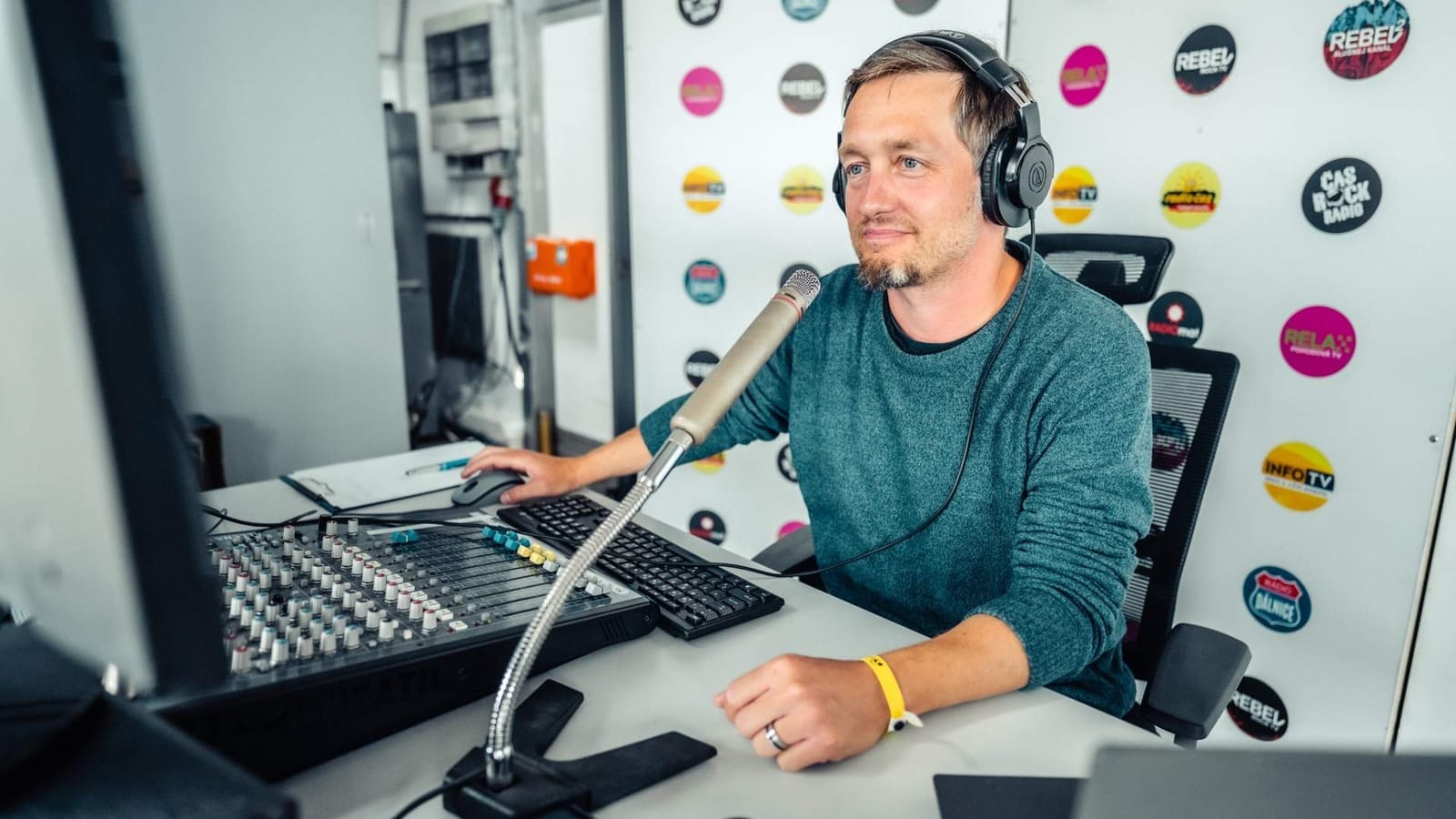 Man wearing headphones sits at a radio mixing desk in front of a wall with station logos including Rebel, Relax and Čas Rock