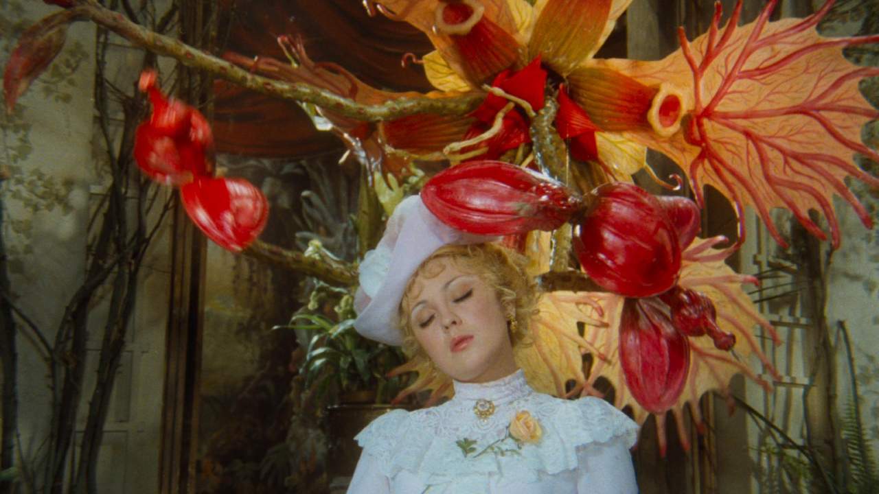 Illustrative still from the film Adéla ještě nevečeřela, with a blonde woman in a white lace dress and hat, eyes closed among giant red and orange fantasy flowers