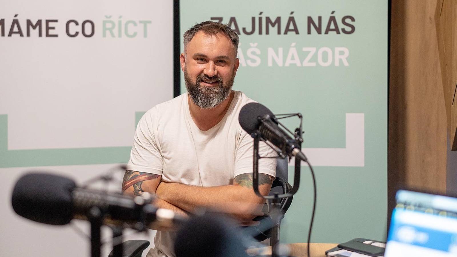 Bearded man in a white T-shirt smiling in a radio studio behind a microphone, with slogans "MÁME CO ŘÍCT" and "ZAJÍMÁ NÁS VÁŠ NÁZOR" on the wall behind him