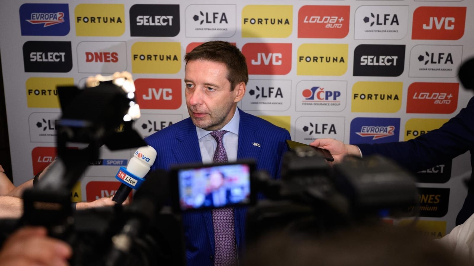 Dušan Svoboda in a blue suit speaking to reporters with microphones and cameras before a backdrop with LFA, Fortuna, JVC, Select and Evropa 2 logos
