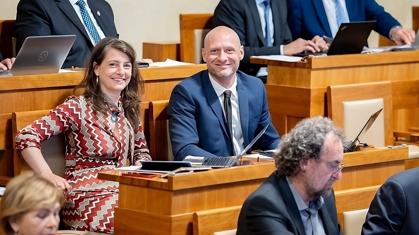 Smiling woman in a patterned dress sits beside Senator Lukáš Wagenknecht in a dark suit at the wooden benches of the Czech Senate chamber, laptops open