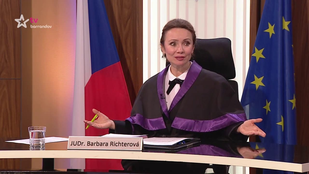 Actress in a judge's robe with purple trim during filming of TV Barrandov's courtroom reality show, nameplate reading "JUDr. Barbara Richterová", Czech and EU flags behind her