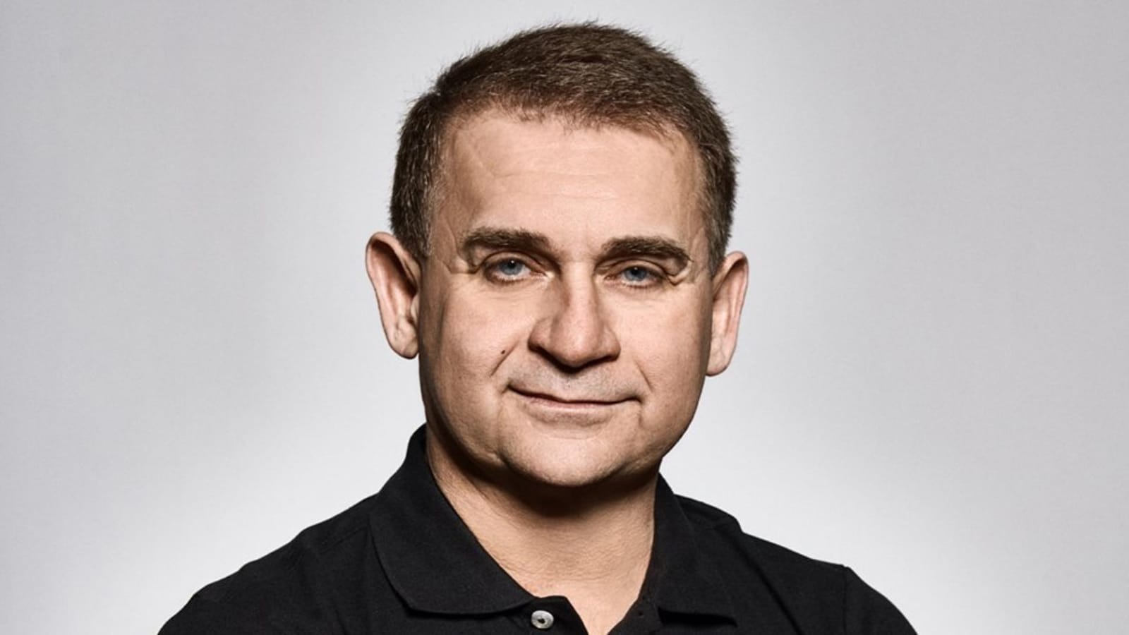 Karel Pražák smiling in a studio portrait, wearing a black polo shirt against a grey background
