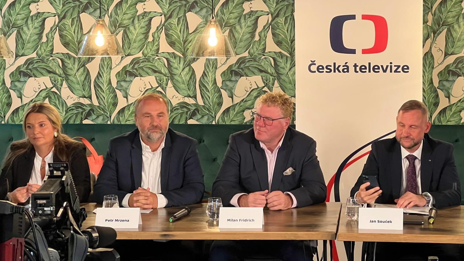 Press conference panel with nameplates reading Petr Mrzena, Milan Fridrich and Jan Souček, beside a Česká televize banner
