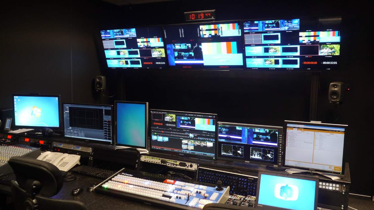 Empty television control room with a wall of monitors, digital clock showing 10:19:44 and a mixing desk with keyboards