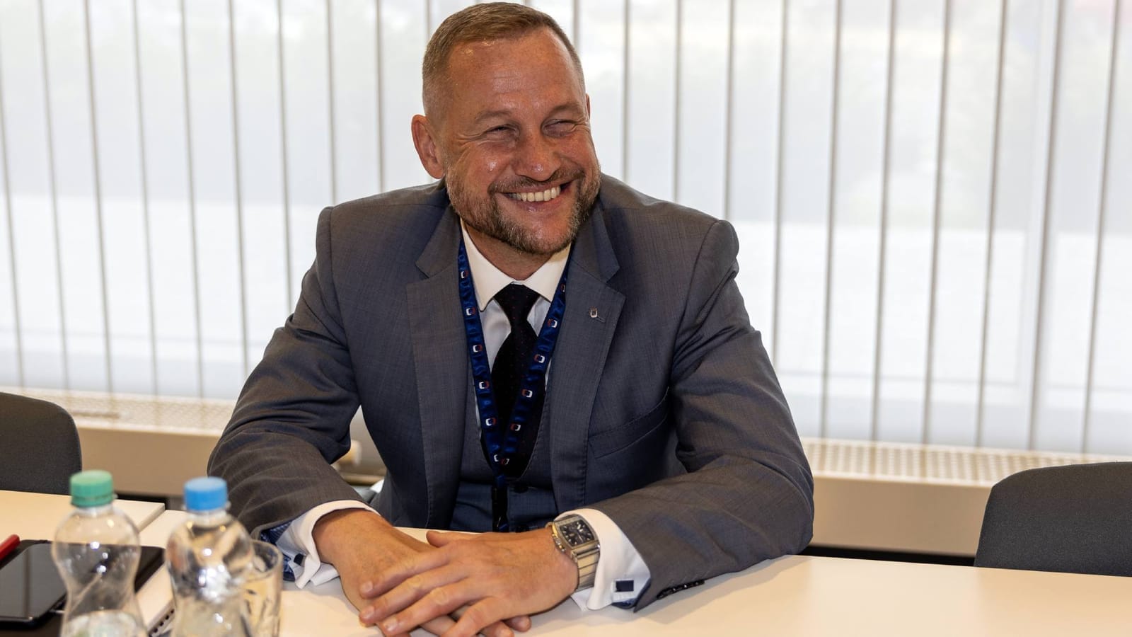 Jan Souček in a grey suit and dark tie, smiling as he sits at a conference table with water bottles