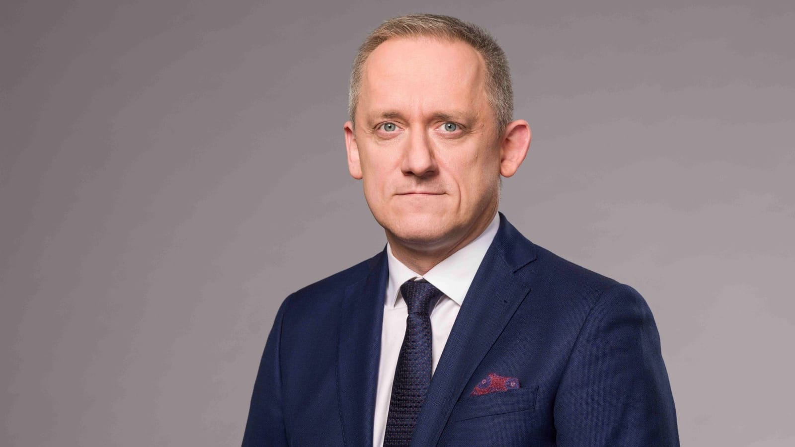 Studio portrait of Michal Kubal in a dark blue suit, white shirt and patterned tie against a grey background