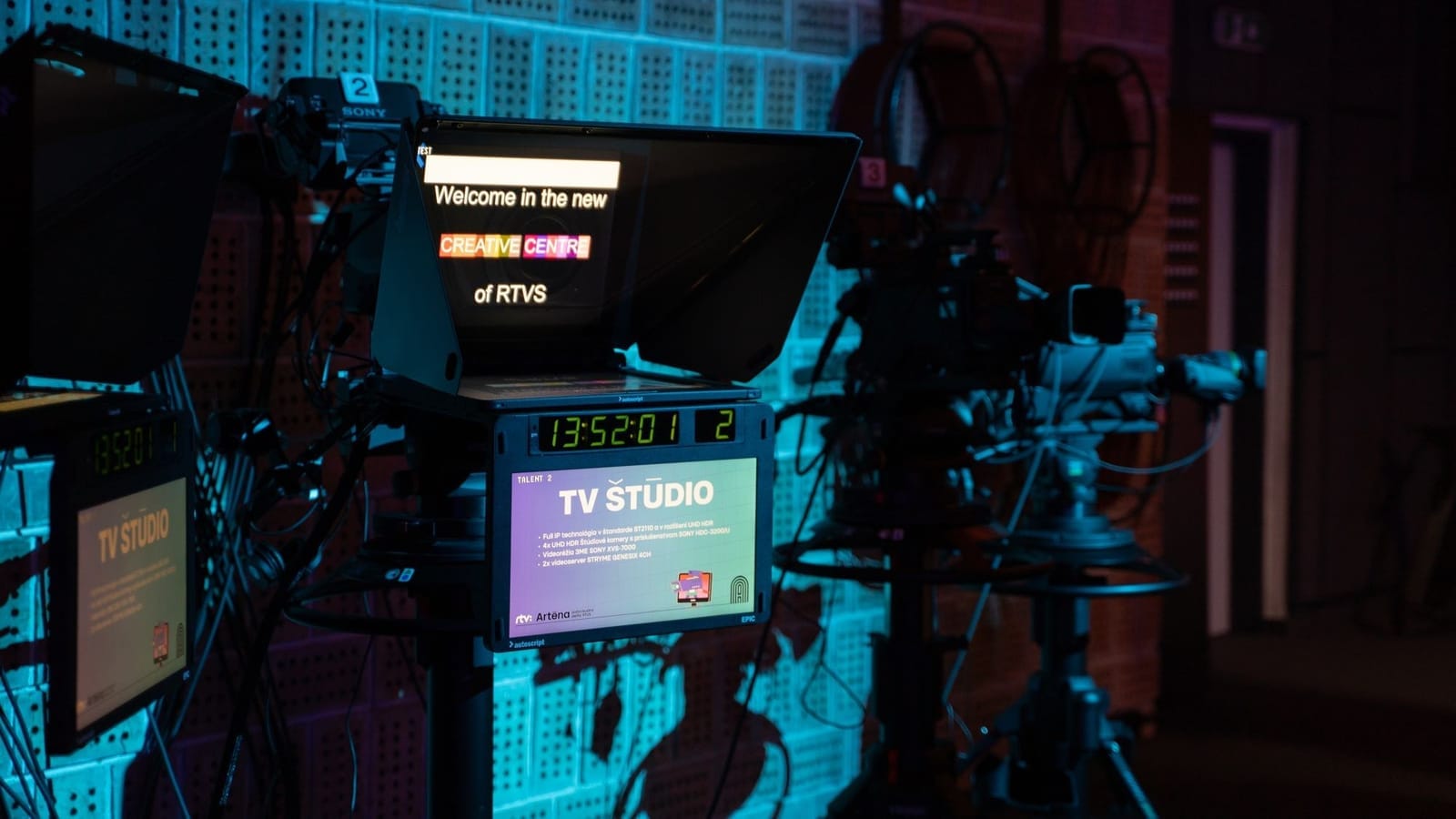 Darkened TV studio with cameras and a teleprompter reading "Welcome in the new CREATIVE CENTRE of RTVS", and a monitor labelled "TV ŠTÚDIO"