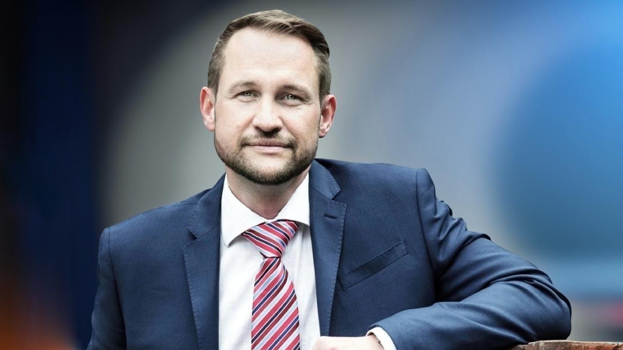 Jan Souček in a dark blue suit, white shirt and red-striped tie, smiling at the camera