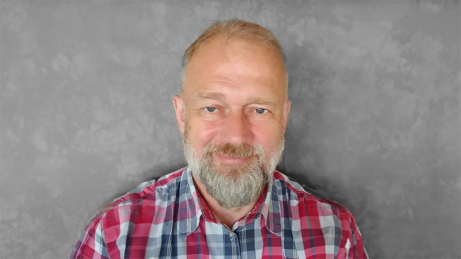 Jan Maxa, smiling with a grey beard and wearing a red and blue checked shirt, against a grey wall