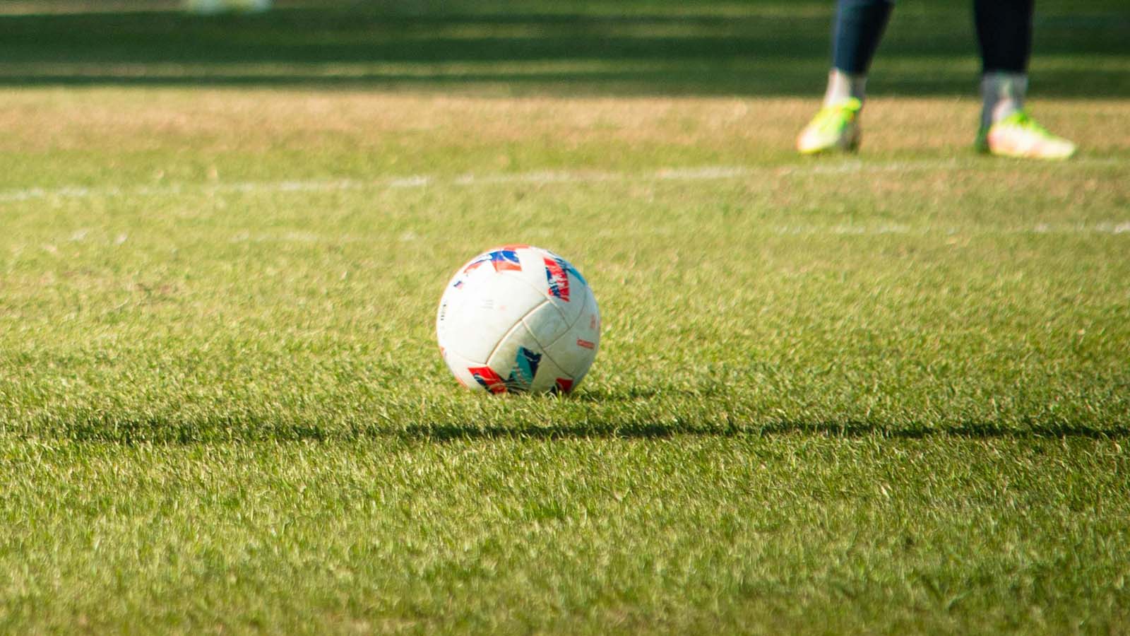 A football lying on a grass pitch, with a player's legs in neon green boots blurred in the background