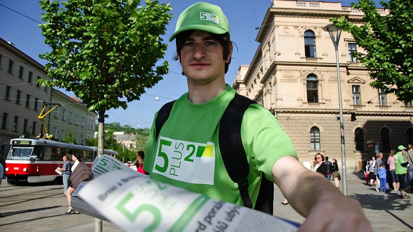 Young man in a green 5plus2 T-shirt and cap handing out free newspapers on a city street with a tram in the background