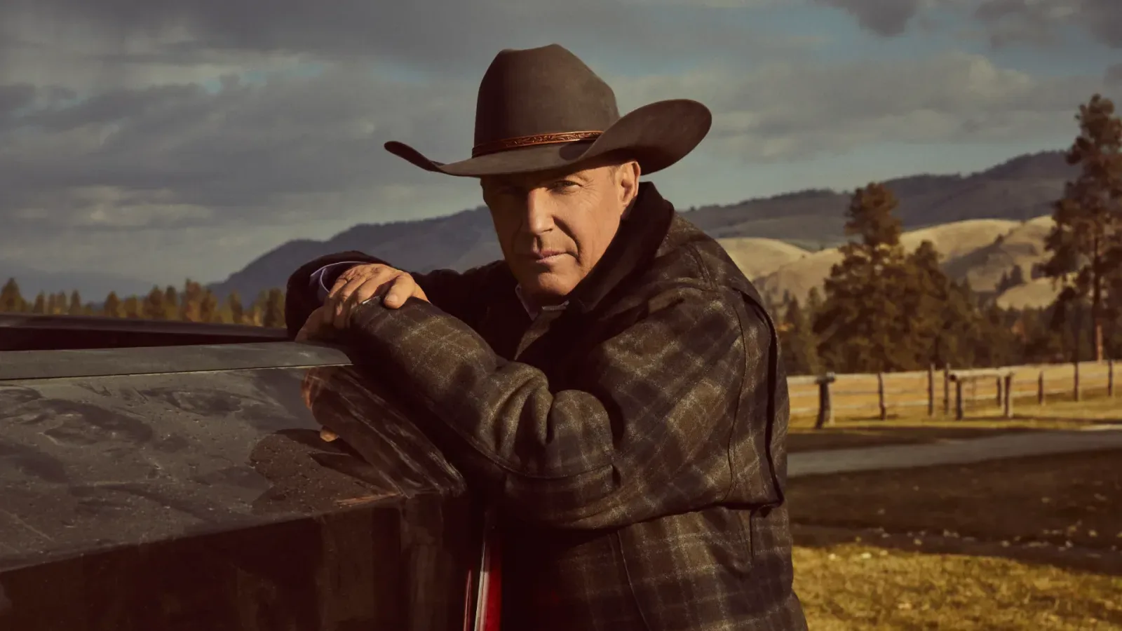Kevin Costner in a cowboy hat and plaid jacket leaning on a pickup truck in a ranch landscape in a scene from the series Yellowstone