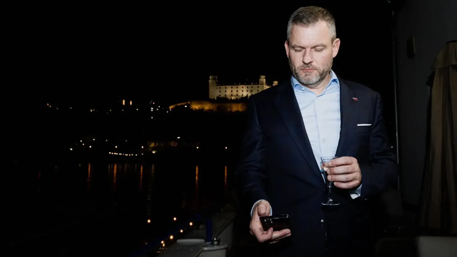 Peter Pellegrini in a dark suit checking his phone with a glass of sparkling wine, illuminated Bratislava Castle behind him at night