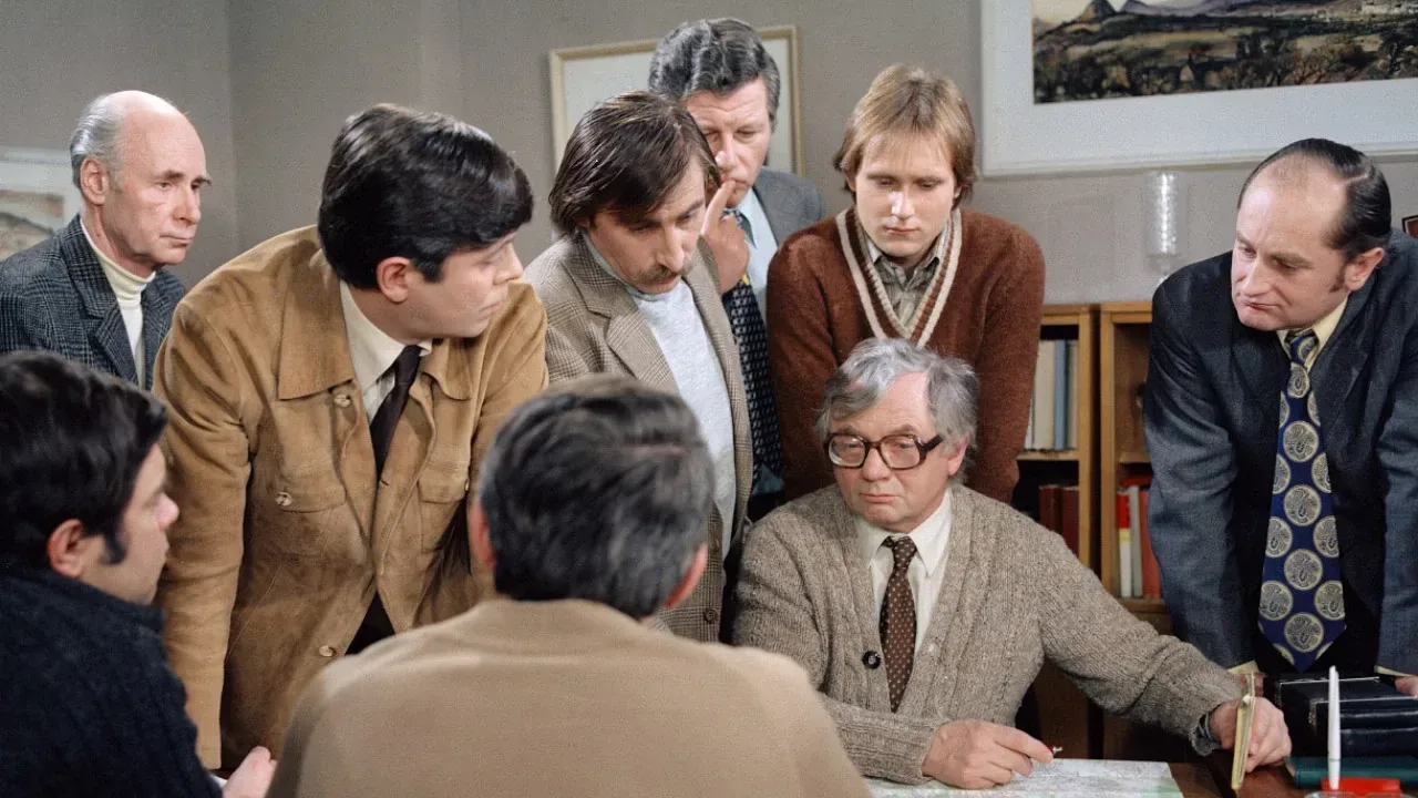 Suited men gathered around a table over papers with a grey-haired man in glasses in a scene from the Czech TV series Malý pitaval z velkého města