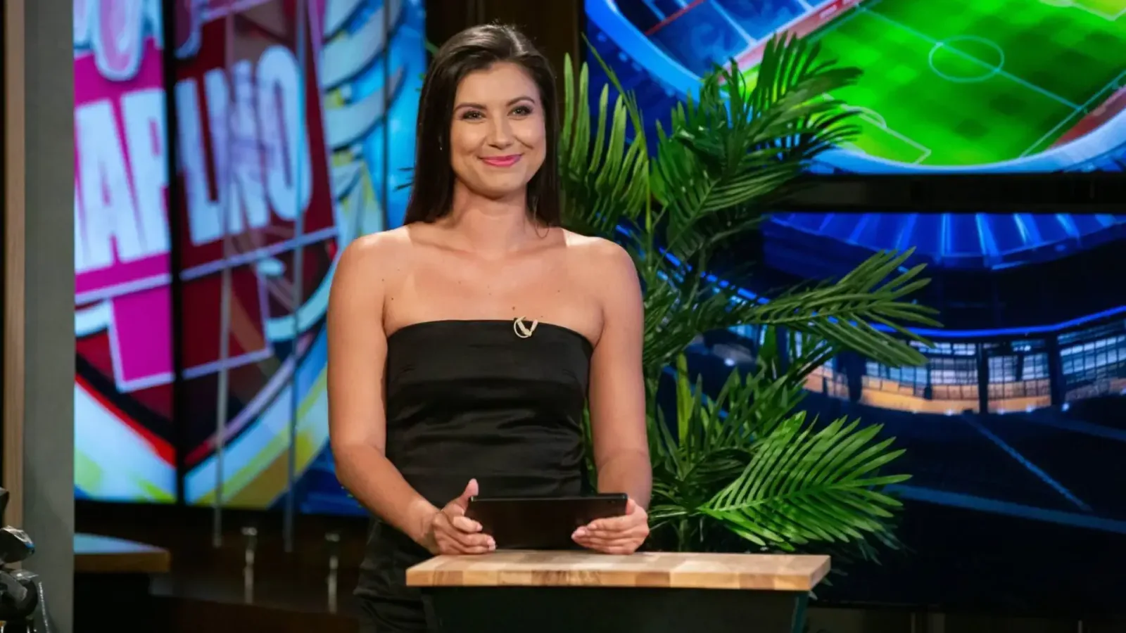 Illustrative shot of an OK TV broadcast, a presenter in a black strapless dress at a wooden lectern before screens with a football stadium graphic