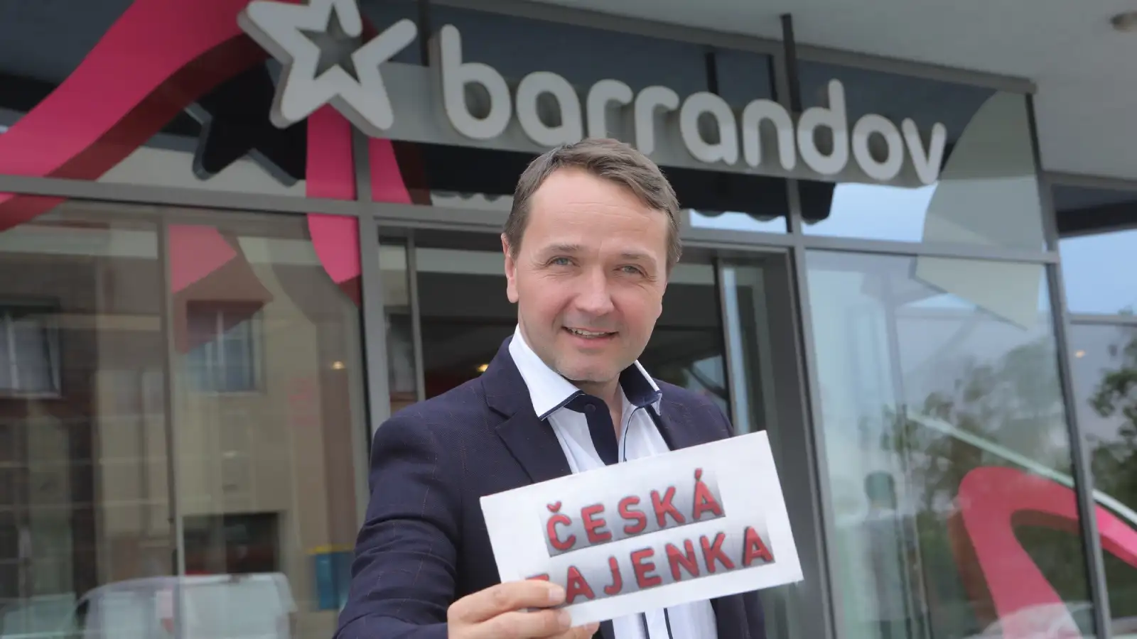 Smiling man in a dark suit holding a "ČESKÁ TAJENKA" card in a shot from the TV Barrandov game show