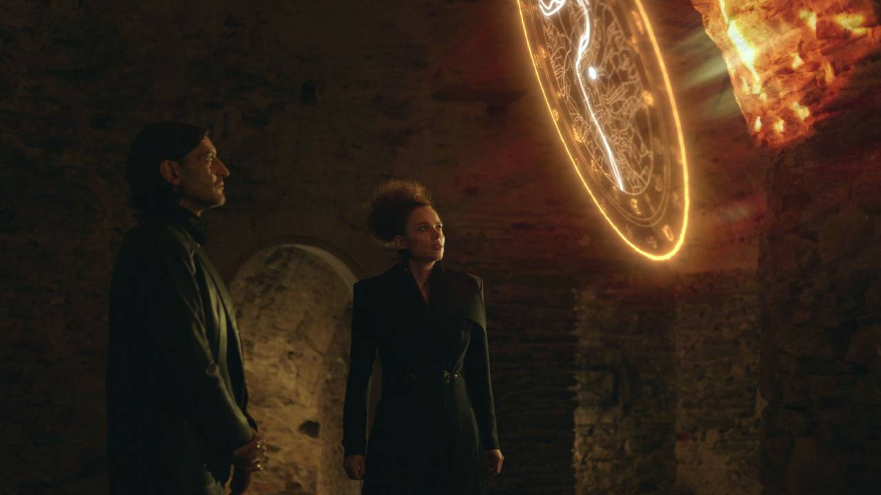 Scene from the series Vědma on Voyo, with a man and woman in dark coats facing a glowing golden portal in a stone vault