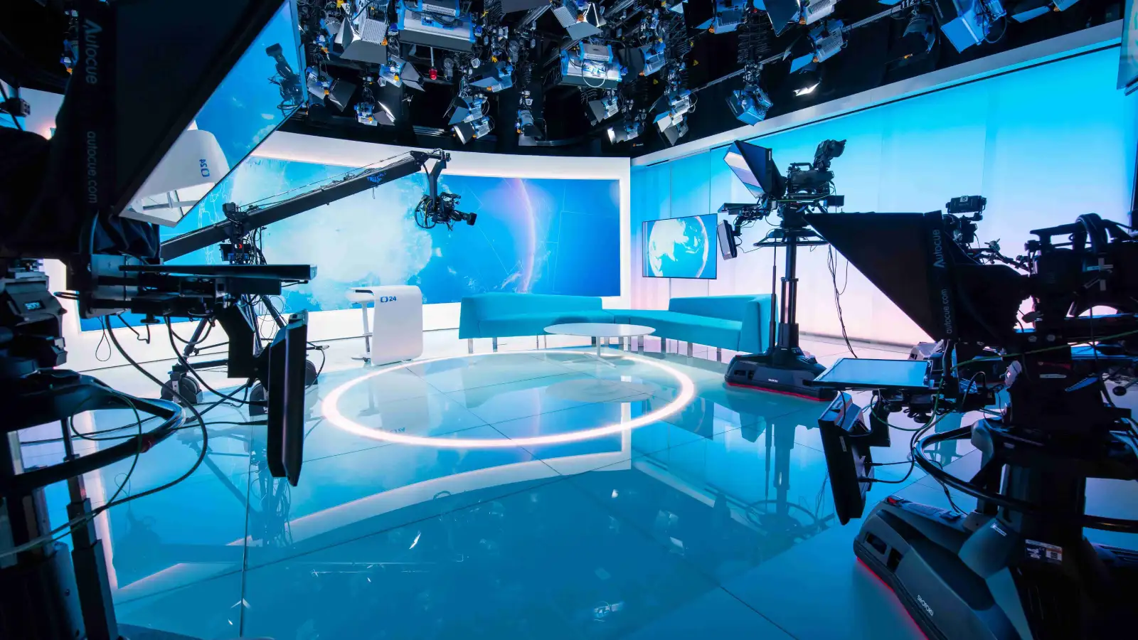 Empty blue-lit television news studio with cameras, a jib arm, teleprompters and a turquoise sofa in front of screens showing a globe