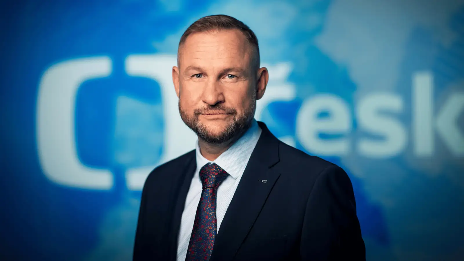 Jan Souček in a dark suit with a patterned tie in front of a blue Czech Television backdrop