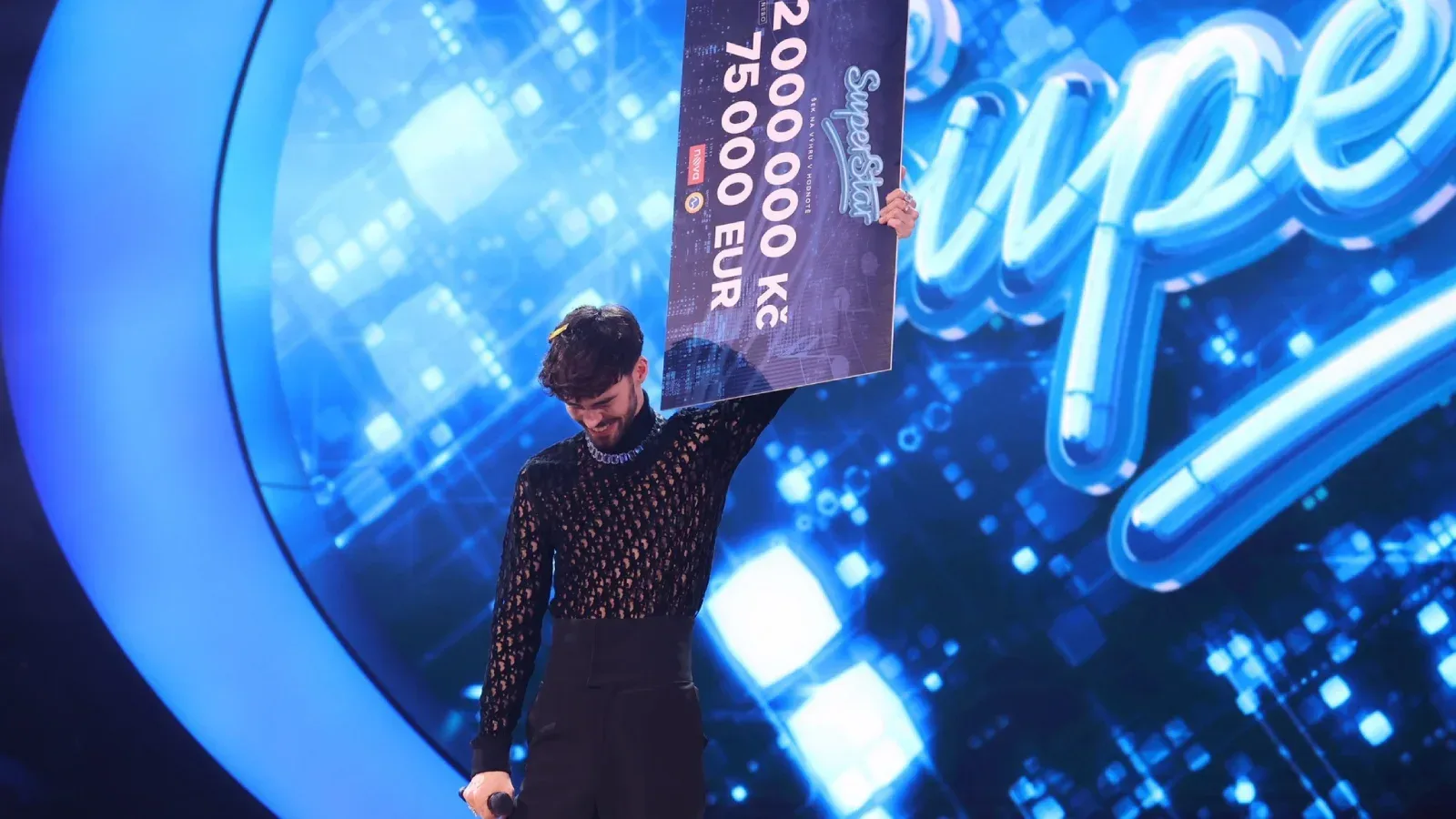 Young man in a black lace top holding up a large cheque reading "2 000 000 Kč / 75 000 EUR" with the Superstar logo on a blue-lit stage