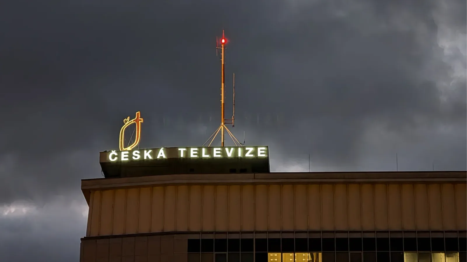 Illuminated "Česká televize" sign and antenna mast on the roof of the broadcaster's building under dark storm clouds