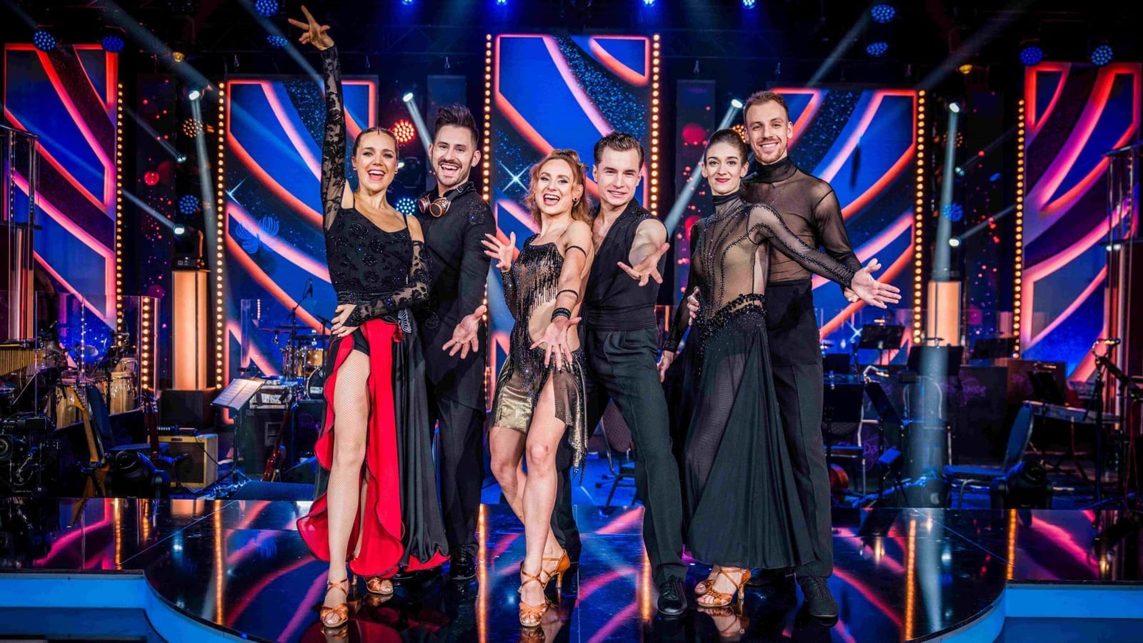 Three dance couples in evening costumes posing on a brightly lit blue and red television stage