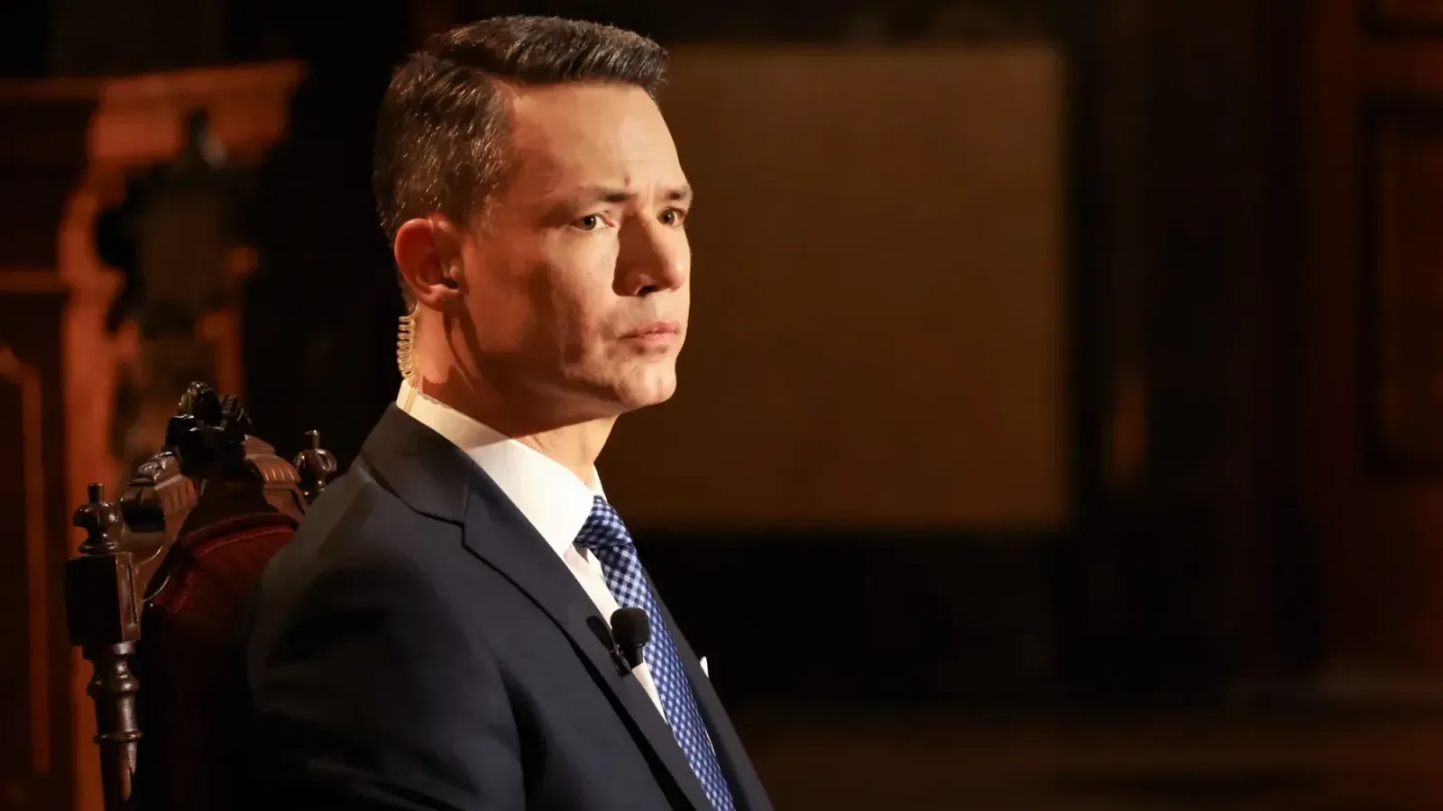 Presenter Martin Řezníček in a dark suit and checked tie, wearing an earpiece and clip-on microphone, seated on the Czech TV studio set where he hosted the presidential candidates' debate