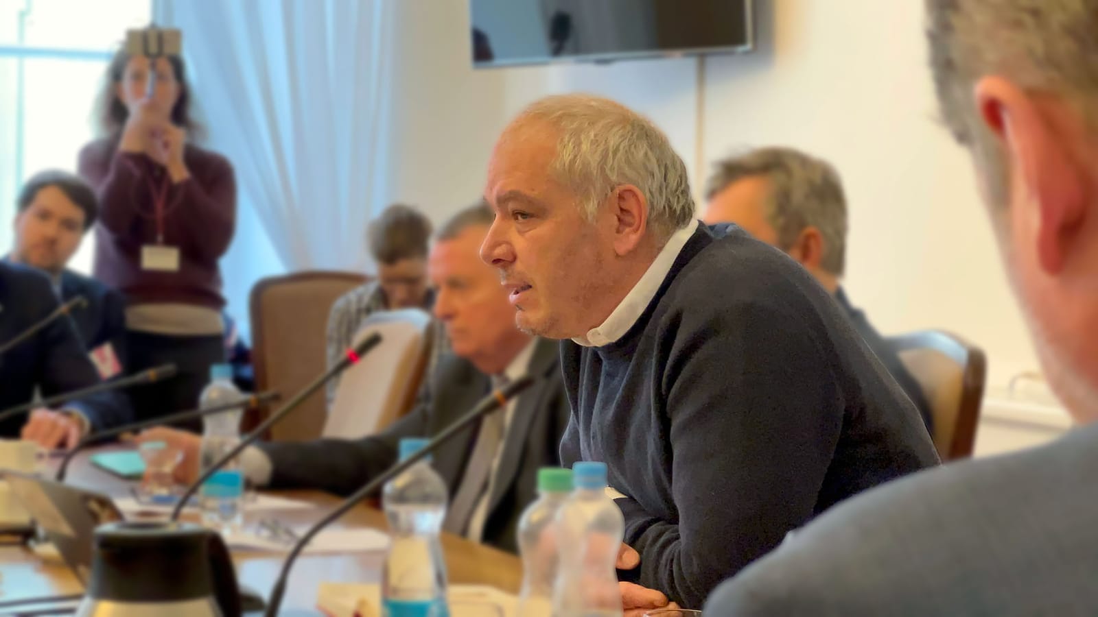 Daniel Köppl in a dark sweater speaking into a microphone at a committee table with seated participants