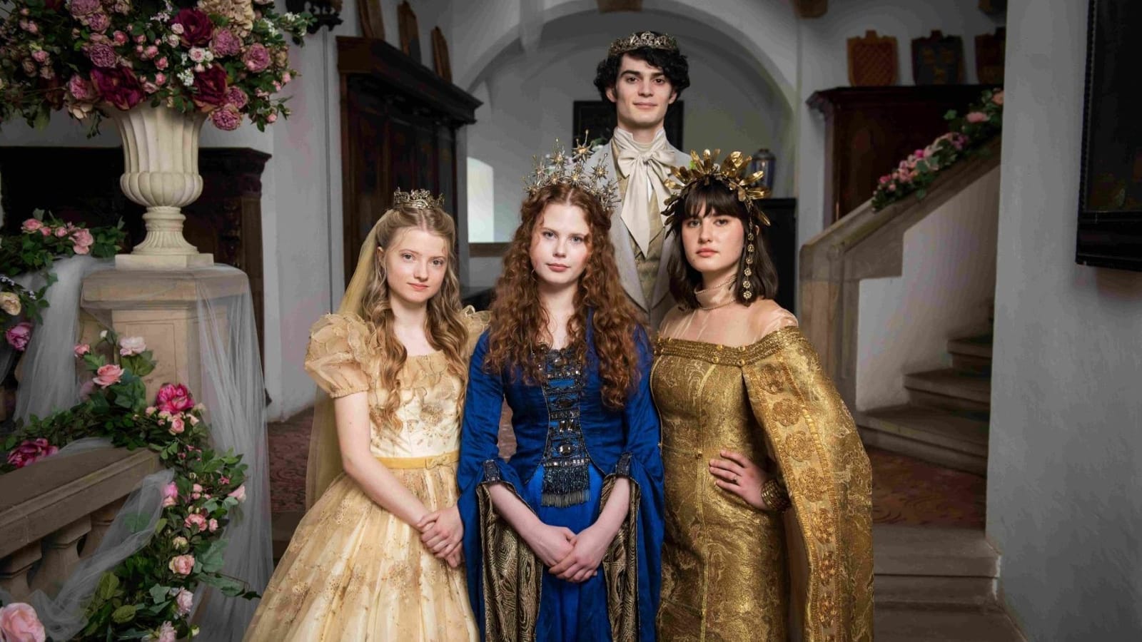Scene from the TV fairy tale Three Princesses, with three young women in ornate historical gowns and golden crowns in a castle hall and a crowned young man behind them
