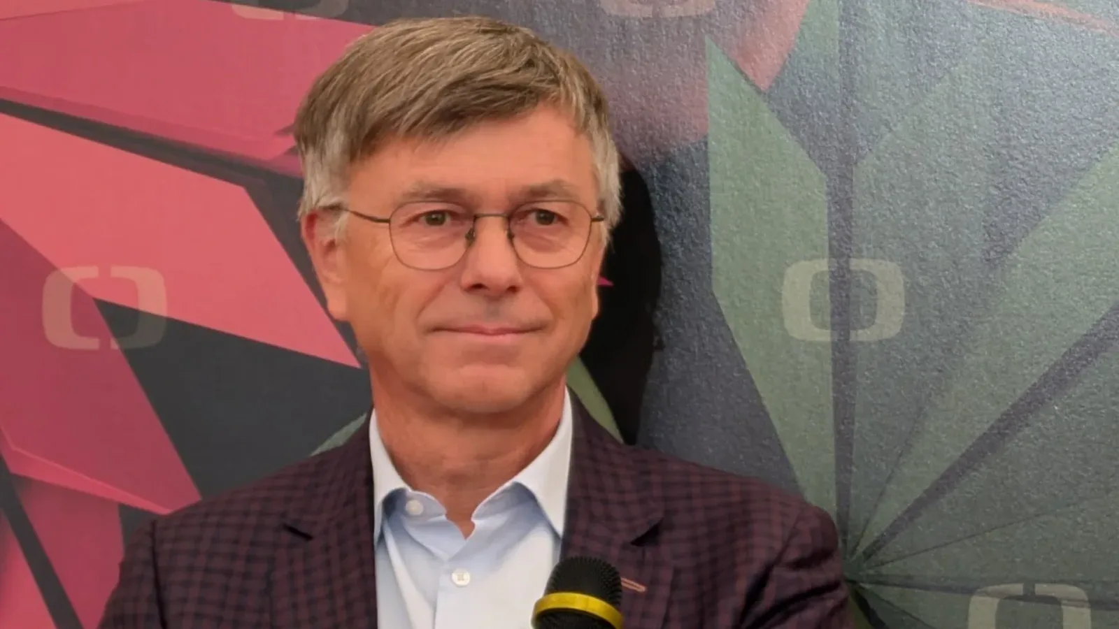 Hynek Chudárek in glasses and a checked jacket holding a microphone in front of a colourful Czech Television backdrop