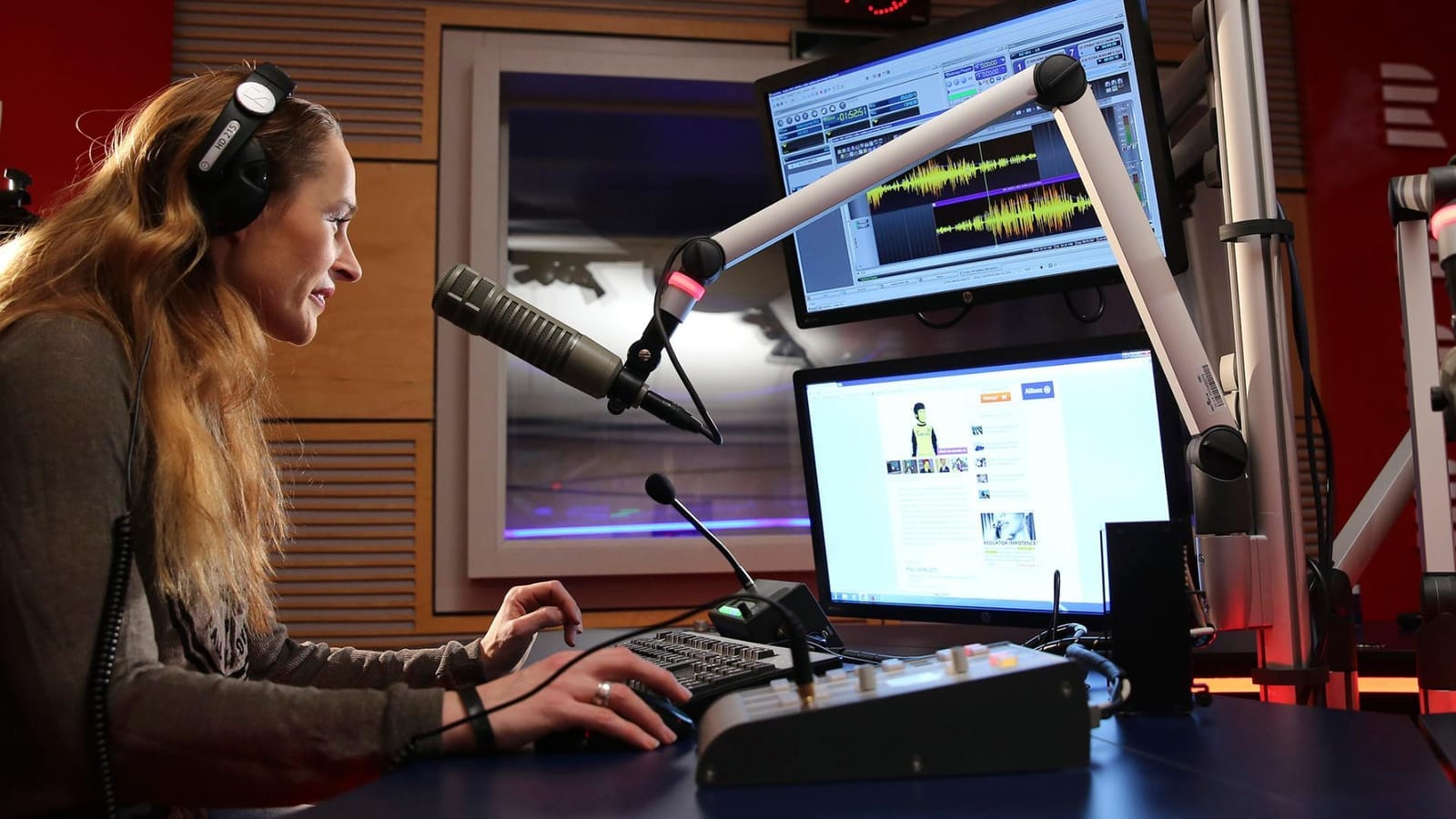 Woman in headphones sits at a radio studio mixing desk in front of a microphone and two screens showing audio waveforms and a website