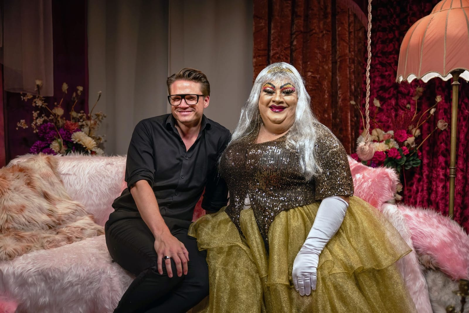A man in a black shirt and glasses sitting on a pink fur sofa beside a drag queen in a gold sequin gown, silver wig and white gloves in a scene from the TV show Chi Chi on the Couch