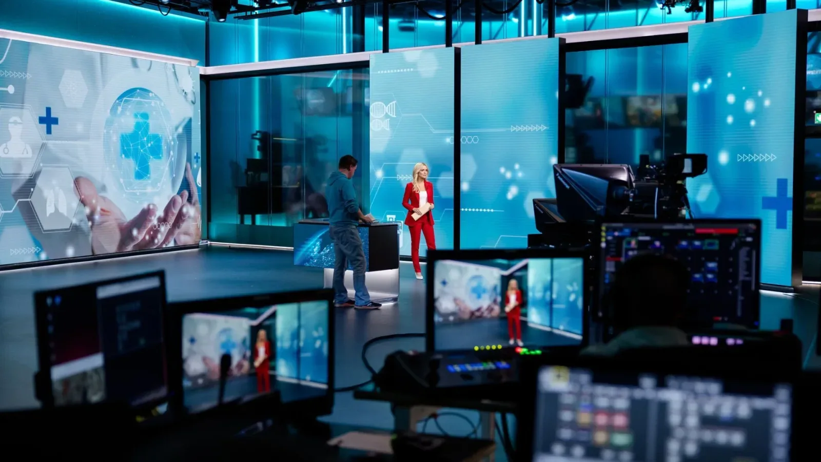 TV news studio with a woman in a red suit at a desk, blue screens, and control-room monitors in the foreground