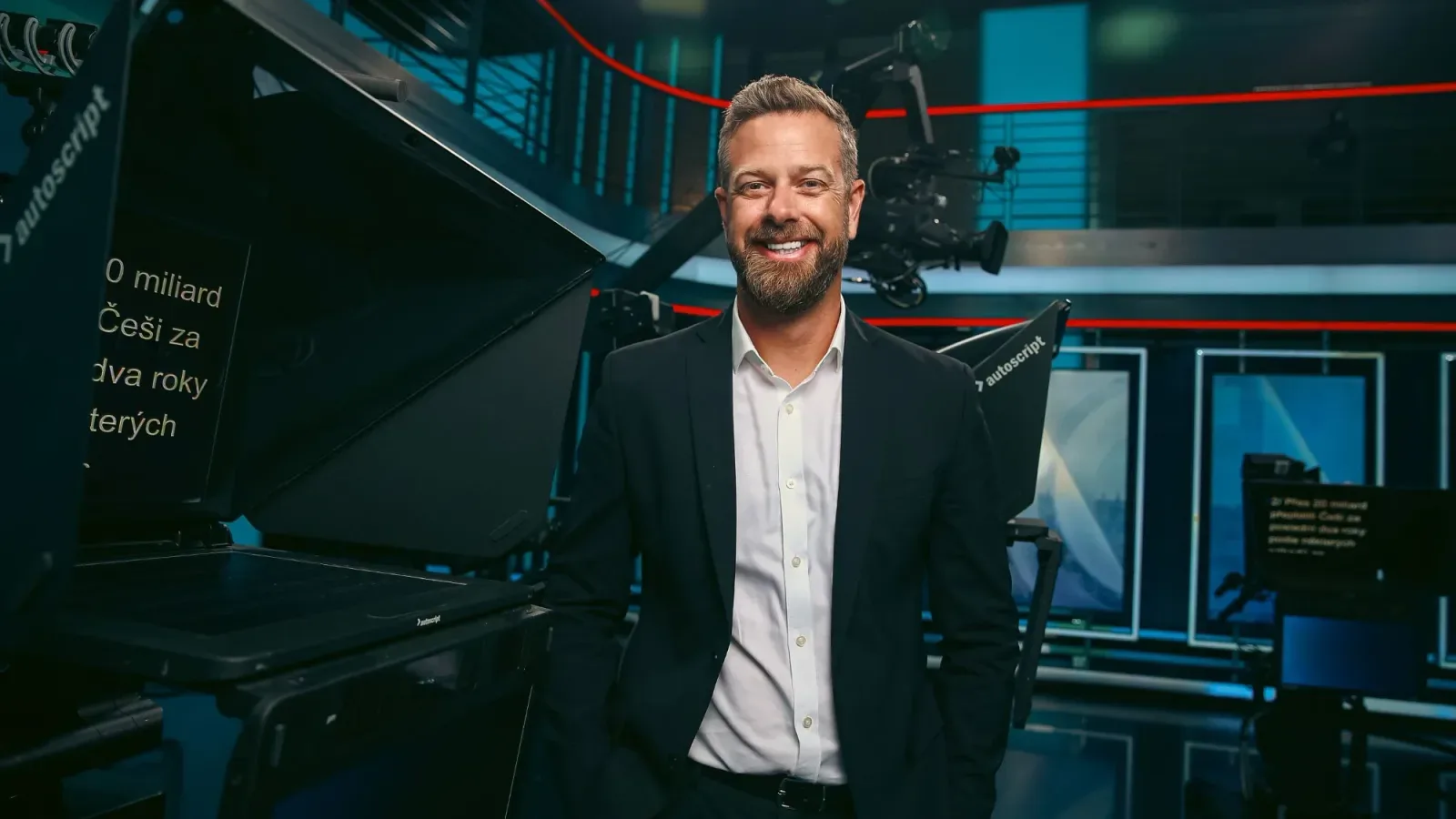 Smiling bearded Lukáš Kubát in a dark suit stands in a TV studio beside a camera with an autocue showing Czech text