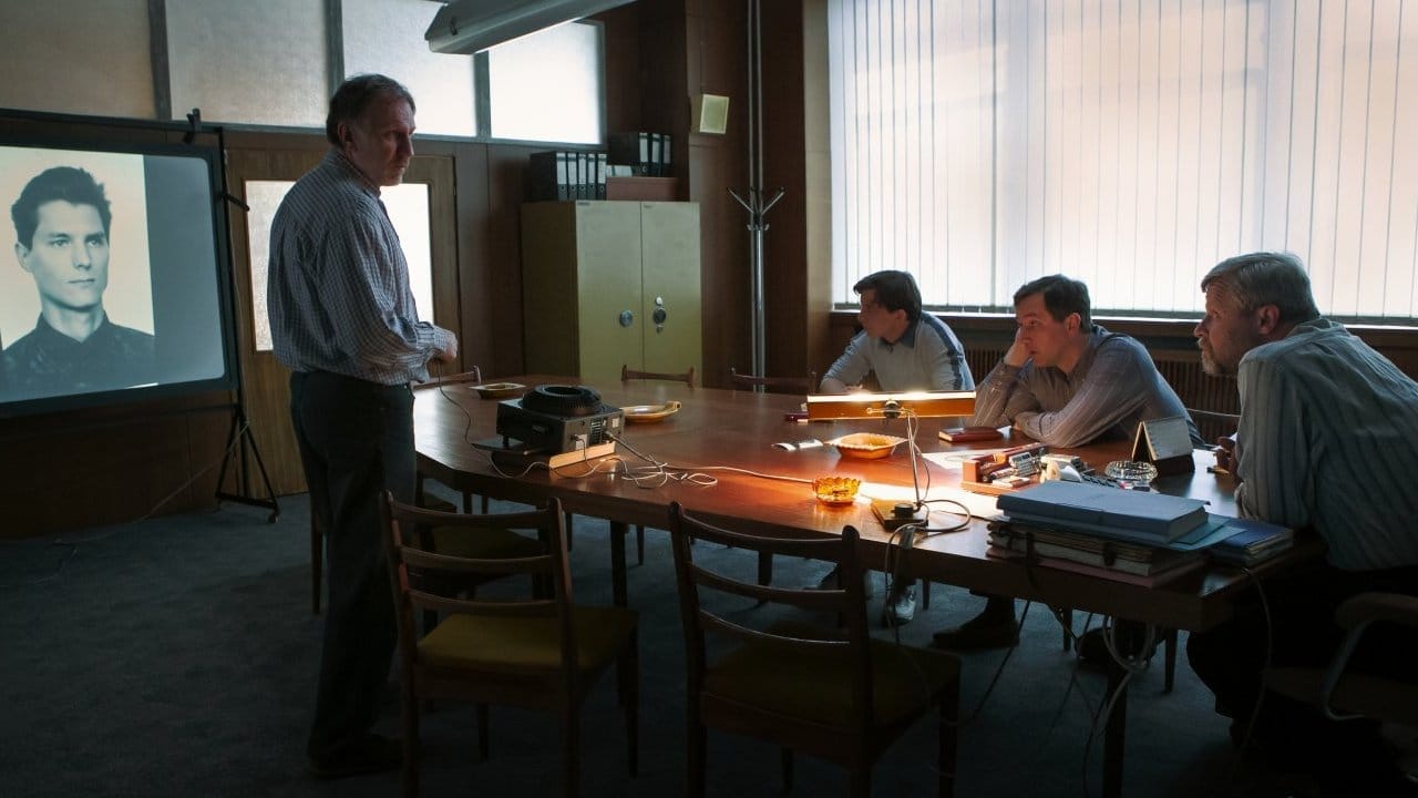 Scene from the TV crime series Devadesátky: a man stands beside a projected black-and-white portrait of a young man as three men sit at a long meeting table in an office