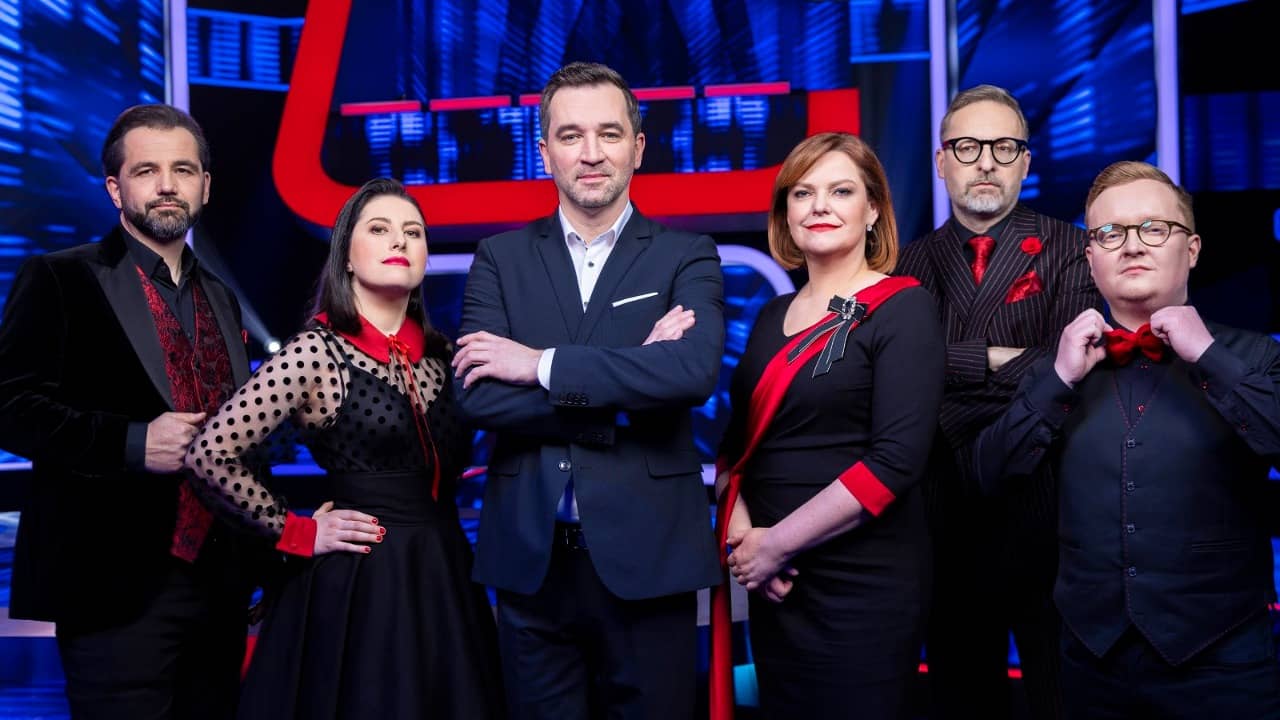 Six contestants in black-and-red outfits surround the suited host in the studio of the TV game show Superlov