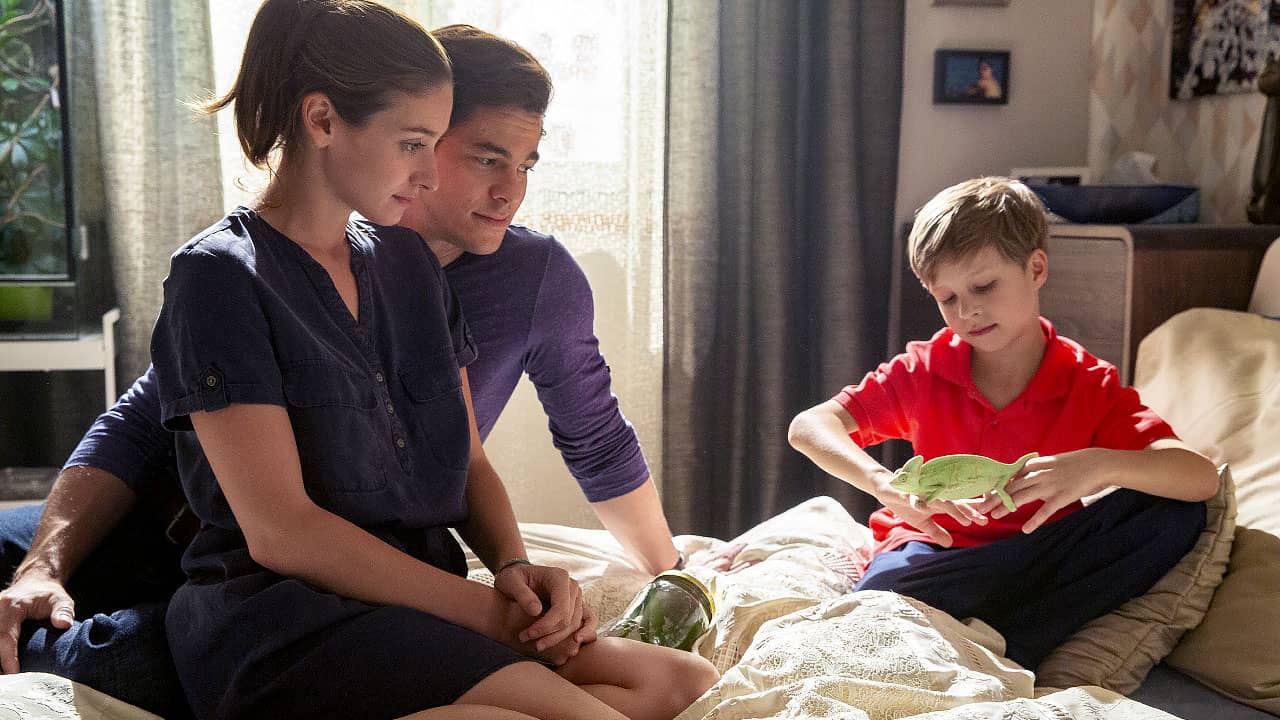 Scene from the TV series Kukačky: a young woman and man sit on a bed watching a boy in a red T-shirt hold a green chameleon