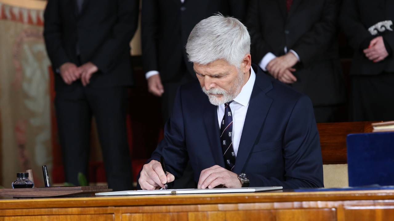 Petr Pavel in a dark suit signing a document at a wooden desk, with people standing behind him