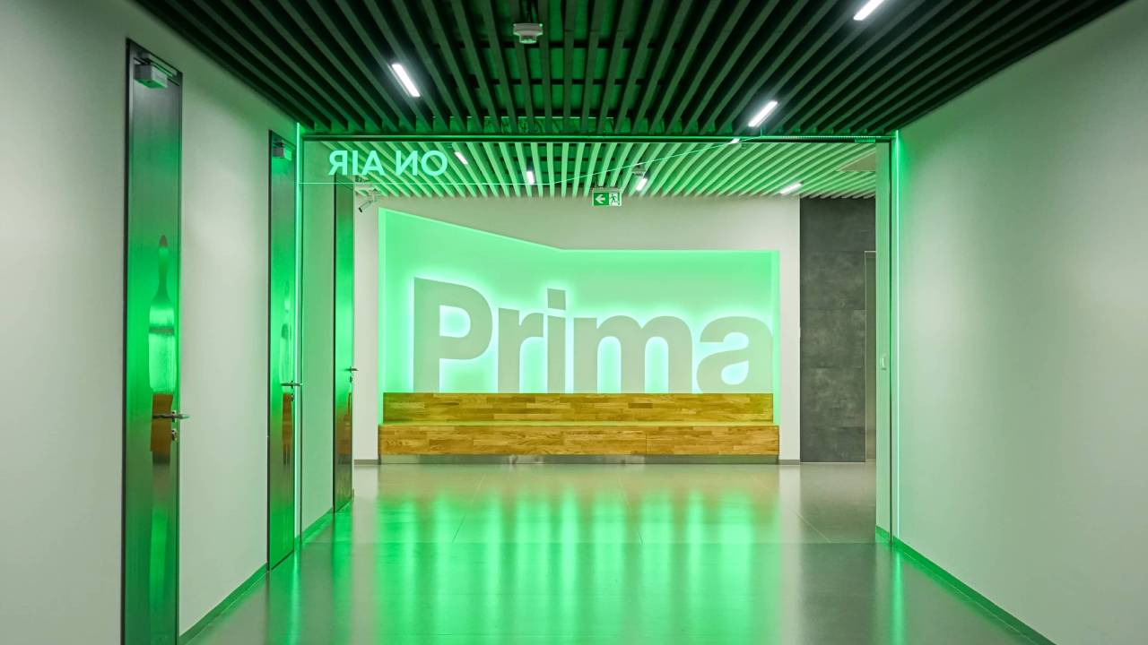 Green-lit corridor leading to a lobby with a large illuminated "Prima" sign above a wooden bench, and an "ON AIR" sign over the glass doors