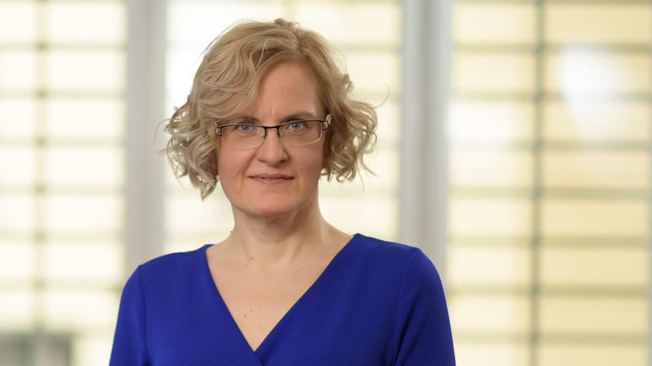 Portrait of Hana Ondryášová, a blonde woman with glasses in a blue V-neck top, in front of window blinds