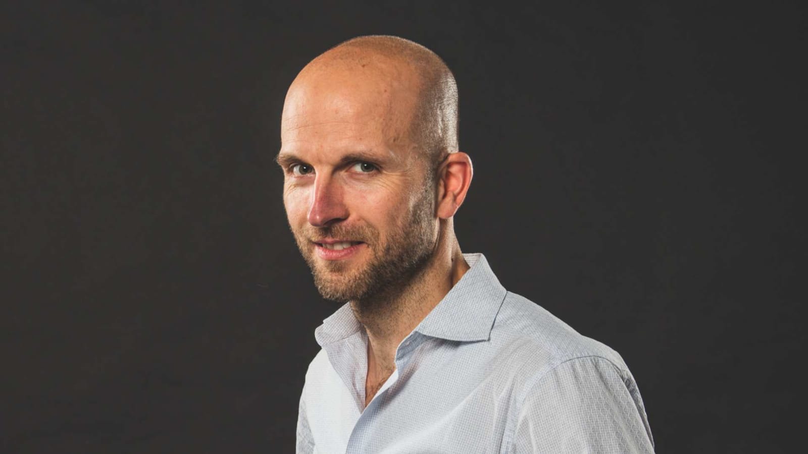 Studio portrait of a bald, bearded man in a light blue shirt against a dark background