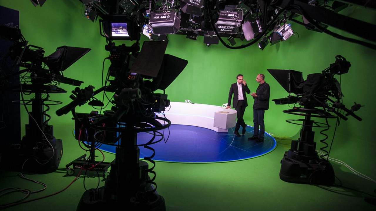 Two men talking in a green-screen television studio surrounded by cameras and lighting rigs