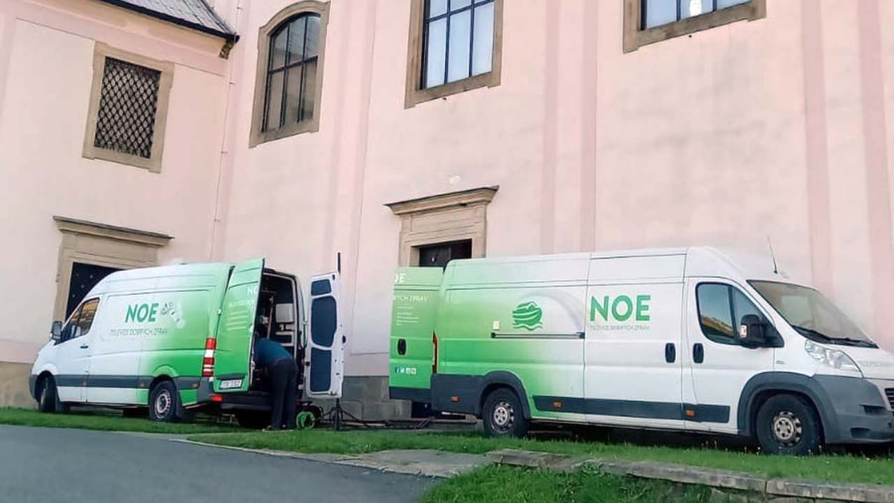 Two white and green vans with the TV Noe logo parked beside a pink church building, one with its rear doors open and a man working inside